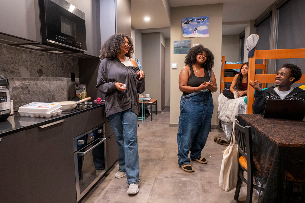 Four people in a modern kitchen and living area with grey cabinets, stainless steel appliances, and a bunk bed.