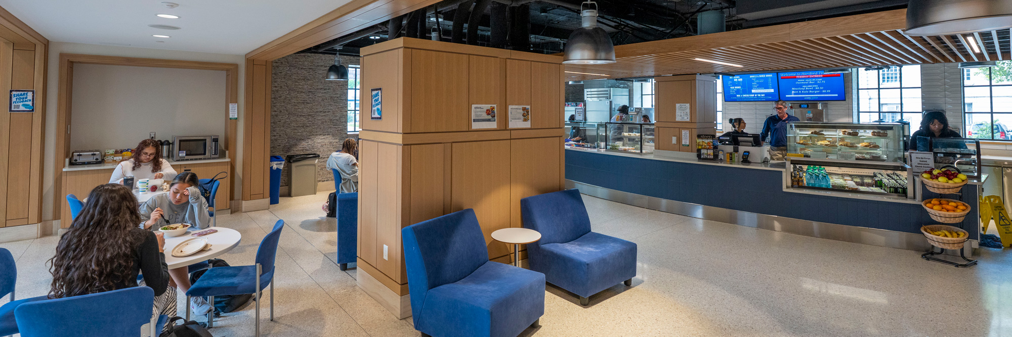 A modern cafeteria with people seated and eating at tables. Blue chairs and couches are arranged around small tables. In the background, a food service counter displays various items, and shelves with fruit baskets are visible on the right. The space features wooden paneling and large windows letting in natural light.