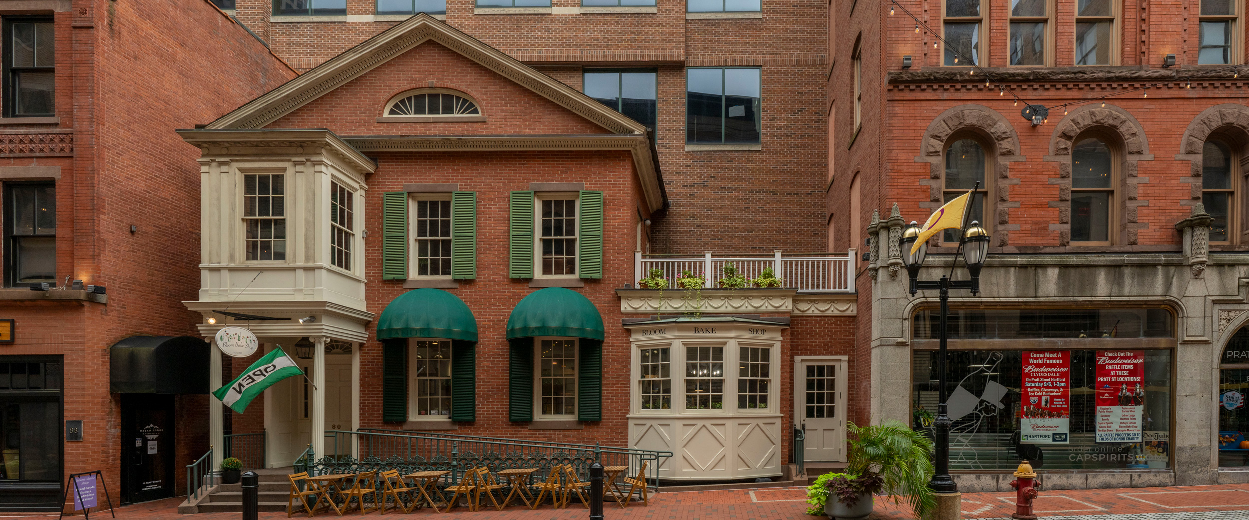 A charming brick building with green shutters and green awnings, featuring a small outdoor seating area with tables and chairs in front. A sign reading "Yard House" hangs above the entrance on the left side. The building is flanked by other brick structures with large windows and decorative architectural details. No people are visible in the scene.
