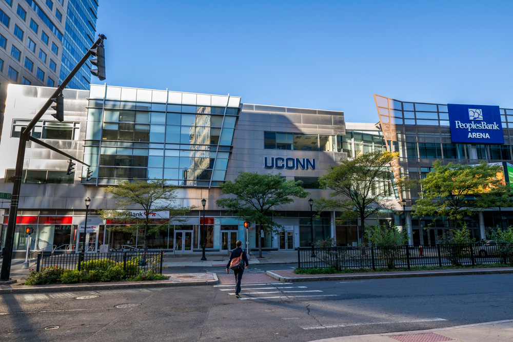 Street view of a modern glass-front building with the word ‘UCONN’ displayed prominently on the facade. To the right, a blue sign reads ‘PeoplesBank Arena.’ Trees line the sidewalk in front of the building, and a person is crossing the street toward the entrance under a clear blue sky.